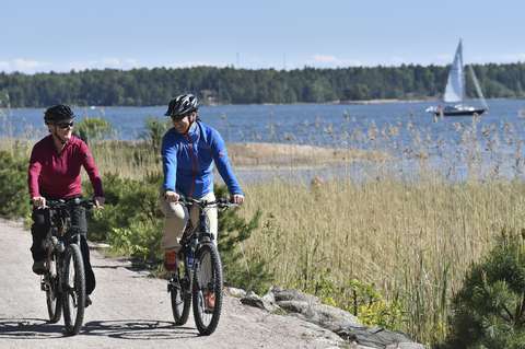 Kaksi pyöräilijää ajavat hiekkatietä pitkin, taustalla näkyy meri ja purjevene. 