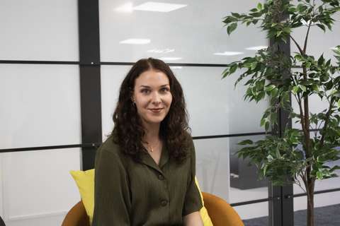 Hanna istuu hymyillen oranssilla nojatuolilla. Taustalla on toimiston lasiseinä ja viherkasvi.