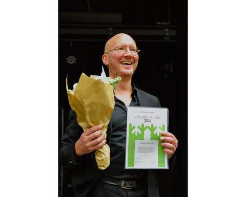 Mies hymyilee kunniakirja ja kukkakimppu kädessä.