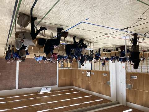 Ihmiset istuvat koulun liikuntasalissa penkeillä kuuntelemassa puhujaa.