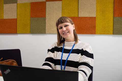 Pia istuu kannettavan tietokoneen ääressä kokoushuoneessa ja hymyilee. Taustalla on värikkäitä akustisia paneeleja.