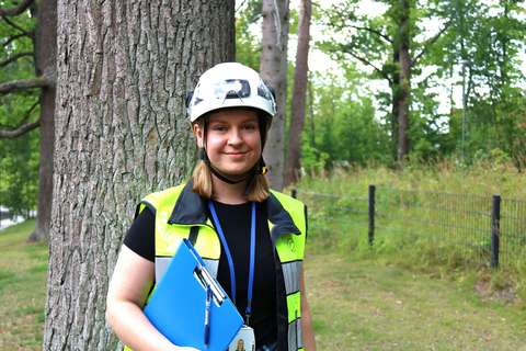 Sonja seisoo puun edessä ja hymyilee. Hänellä on kypärä päässä, työliivi yllään ja sininen kansio kädessä. Taustalla on aita ja kasvillisuutta.