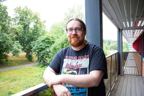 Ukko seisoo parvekkeella. Hän nojaa kaiteeseen ja hymyilee. Taustalla on viherkasvustoa.