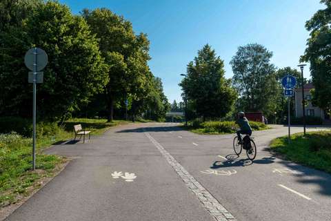 Gångväg och cykelväg som omges av trafikmärken, bänk och träd. En cyklist cyklar på cykelfältet. 