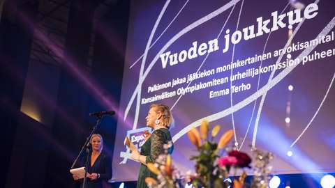 The picture shows the main stage of the Sportsgala of 2024. Two women are standing on the stage. There are some flowers in front of the women. On the wall is written Vuoden joukkue.
