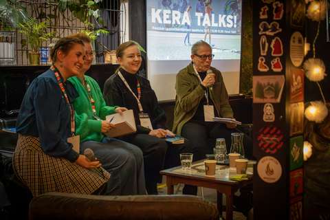 Neljä henkilöä istuu lavalla paneelikeskustelussa Kera Talks! -tapahtumassa. Taustalla näkyy tapahtuman logo valkokankaalla, ja tilan sisustus on rento ja olohuonemainen, sisältäen viherkasveja ja koristevaloja. Paneelissa olevat henkilöt pitelevät mikrofoneja ja muistikirjoja, ja keskustelun tunnelma vaikuttaa keskittyneeltä ja lämpimältä.