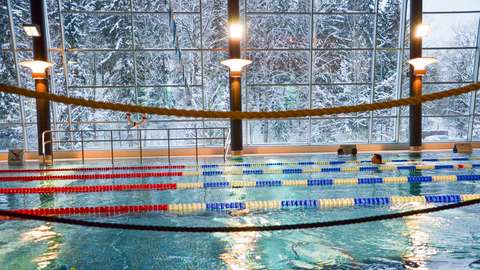 The picture shows the big swimming pool in the Keski-Espoo swimming hall. In the background you can see snowy trees through a window.