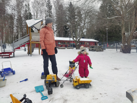 Lapsi leikkii lumisen asukaspuiston pihalla nukenrattailla. Äiti seisoo vieressä.