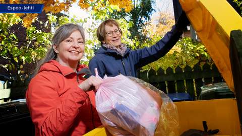 Reetta Jänis is looking at the camera and placing a garbage bag into the trash bin, while Minna-Leena Ahola is holding the lid open