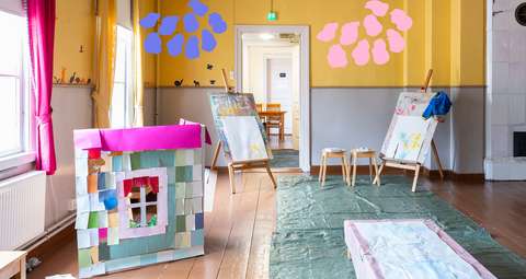 Huoneessa maalaustelineet sekä lattialle laitettu pitkä paperi maalausta varten. Värikäs leikki seinäke on huoneessa.