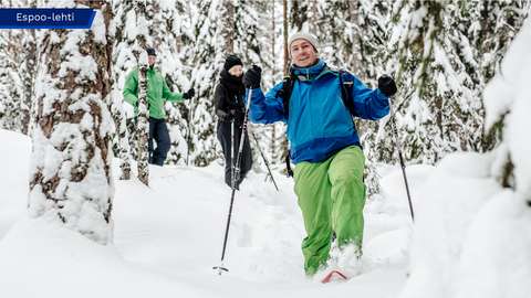 A person is walking in a snowy forest wearing snowshoes and smiling towards the camera.