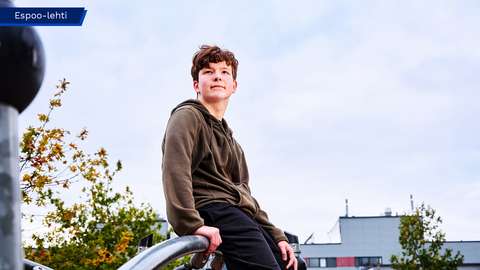Pietu is sitting on top of a climbing frame and looking towards the camera, with Vanttila School in the background.