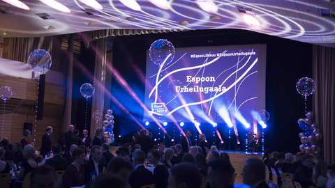 The picture shows the scene of the Espoo Sports Gala from a previous year. There are people in front of the scene and in the background you can see lights in different colors.