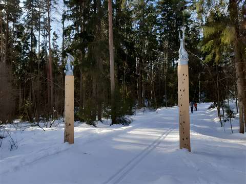 Kuvassa keskuspuistossa sijaitseva portti, jossa kahden puupylvään päällä metalliset jänikset. Portin läpi menee latu aurinkoisessa säässä.