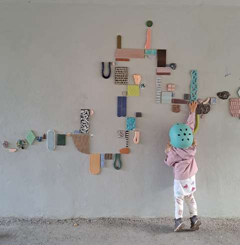 Seinällä on erilaisista irrallisista kappaleista koostuva taideteos, jossa on erilaisia värejä ja muotoja. Lapsi seisoo seinän edessä selin kameraan ja kurottautuu tutkimaan teosta.  Lapsella on päällään vaaleansävyiset vaatteet ja päässä mintunvihreä pyöräilykypärä.