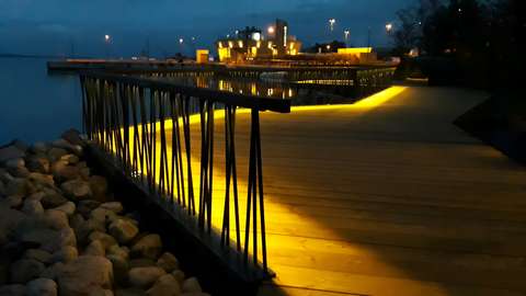 En gångbana och utsiktsterrass i trä alldeles intill havsstranden i Nokkala. Terassen kantas av ett staket av metallspjälor och är försett med en ledstång i trä. Under ledstången finns LED-lampor som belyser träterrassen vackert.