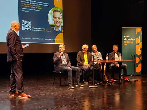 Picture from the “Taloyhtiöiden energiailta” seminar held at the Tapiola Cultural Centre. In the photo, four people are sitting on stage talking. The event host is standing on the left side of the photo.