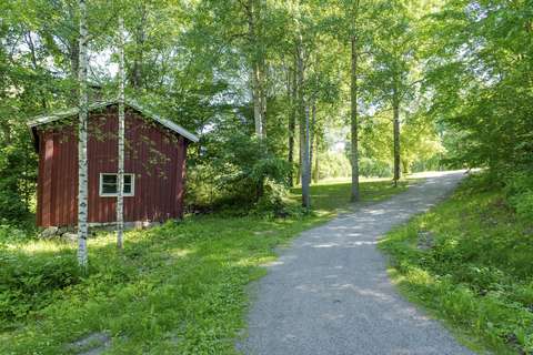 Kuvan oikeassa reunassa on puistoraitti, joka kulkee lehtipuiden välissä. Kuvan vasemmassa reunassa on vanha punainen rakennus.