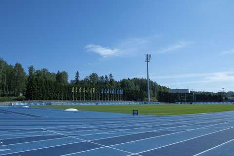 Kuvassa on Leppävaaran yleisurheilukenttä. Etualalla näkyy sinistä juoksurataa ja juoksuradan keskellä oleva nurmialue. Taustalla näkyy kentän korkea valaisinmasto, rivi lippuja sekä metsää.