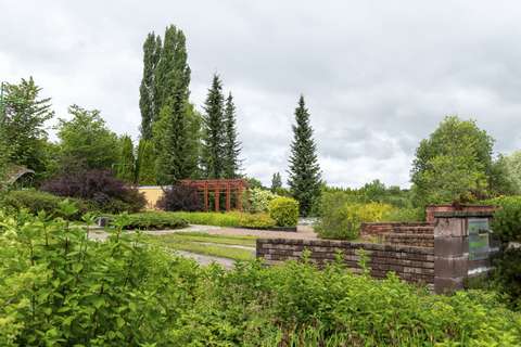 Kuvassa on näyttelypuiston moninaista puutarhakasvillisuutta ja tukimuurirakennelma. Taustalla näkyy pergolarakennelma. 