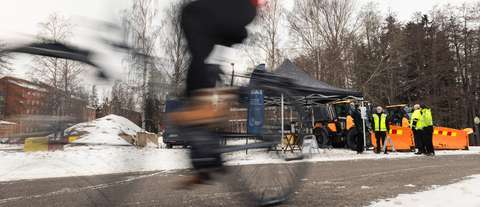 Pyörätie, jossa pyöräilijä ajaa vauhdikkaasti. Takana näkyy harjasuolauskoneita ja ihmisiä seisomassa teltan vieressä talvisessa maisemassa.