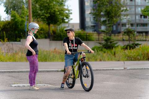 Kaksi ihmistä kesällä asfalttipihalla. Toinen istuu pyörän satulassa ja toinen seisoo.
