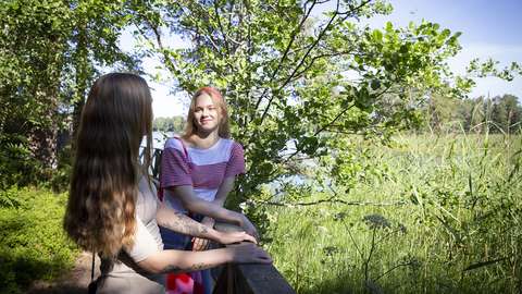 Kuvassa kaksi nuorta naista seisoo kaiteen luona kaislikon ja metsän välissä. Kuvassa on kesä.