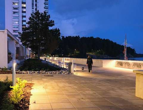 På bilden syns Strandpromenaden i Stensvik i kvällsbelysning. En människa promenerar med en liten hund. I bakgrunden syns ett högt tornhus.
