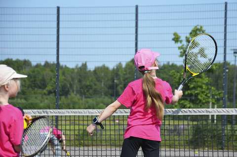 Bilden visar två flickor som spelar tennis på ett uteplan. I bakgrunden finns grön natur.