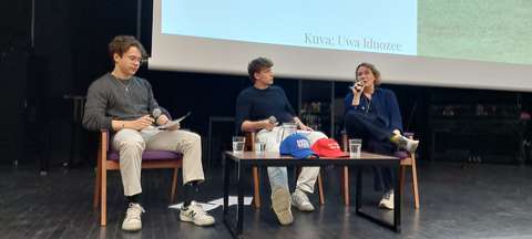 Haastateltava ja kaksi haastattelijaa istuvat tuoleilla auditorion lavalla.