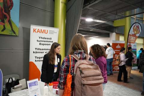 KAMUlaiset Hello Espoo tapahtuamassa seisomassa ständin pöydän takana ja juttelemassa kahden kuvassa selvin olevan henkilön kanssa.