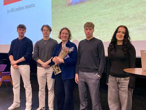 Luennoitsija seisoo auditorion lavalla kukat kädessä neljän muun henkilön kanssa.