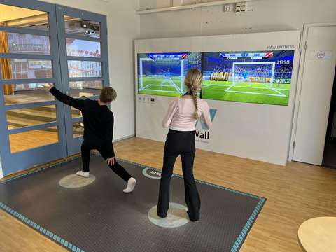 Kaksi oppilasta pelaamassa liikuntapeliseinällä jalkapalloa.