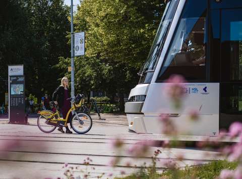 Polkupyräilijä seisoo pysäkillä. Taustalla puita ja etualalla raitiovaunun keula.