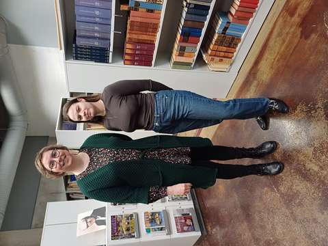 Kaksi henkilöä, Kirsi ja Anna, seisovat kirjahyllyn edessä poseeraten.