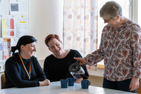Kaksi hymyilevää työntekijää istuu kahvipöydässä ja yksi kaataa heille kahvia.