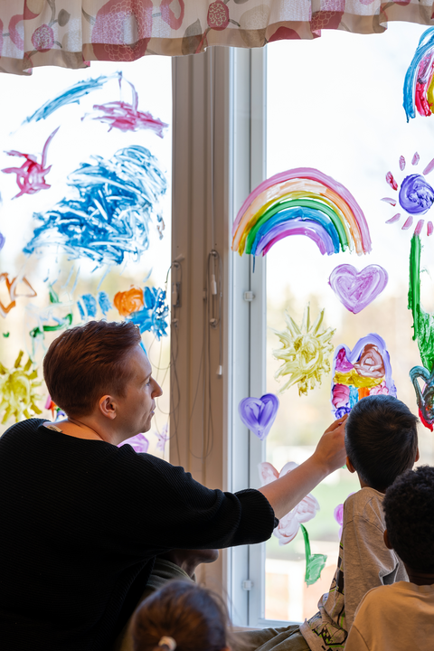 Opettaja ja lapset maalaavat ikkunaan värikkäitä kuvia kuten sateenkaaria.