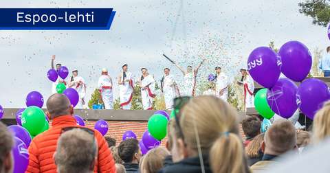 Studenter klädda i vita togor firar på ett tak, medan publiken på evenemanget håller i ballonger.