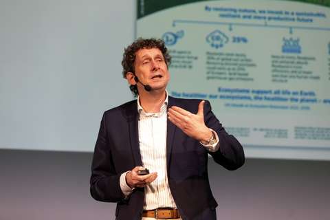 En prydligt klädd person talar i en mikrofon framför en stor diabildskärm.