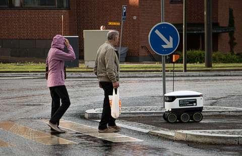 Suojatietä ovat ylittämässä kaksi jalankulkijaa ja ostoksia kuljettava robotti.