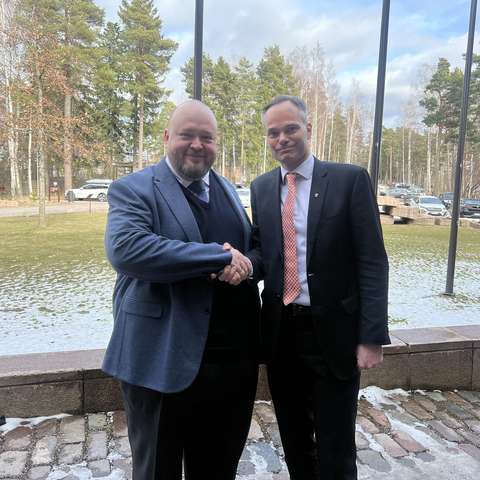 Kaksi pukuihin pukeutunutta miestä katsoo kameraan ja kättelee toisiaan.