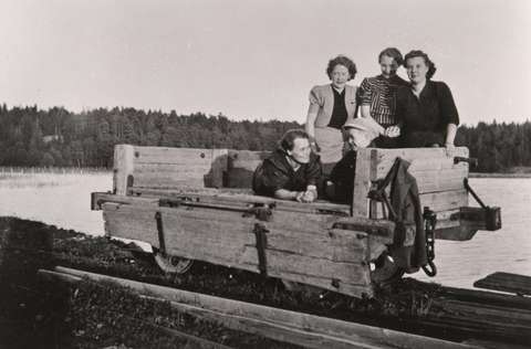 mustavalkoisessa valokuvassa merenlahti ja raiteella kulkeva puinen vaunu, jonka kyydissä on neljä naista ja lapsi.