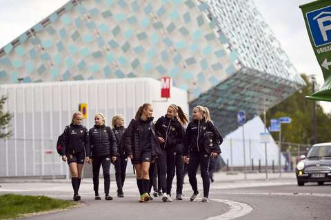 Joukko nuoria urheilijoita kävelee kadulla. Taustalla näkyy vino, kuutiomainen rakennus, joka on metroasema.