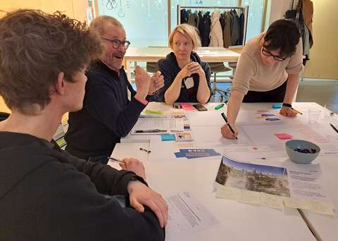 Valokuvassa neljä eri-ikäistä henkilöä istuu pöydän ääressä. Yksi kirjoittaa suurelle paperille, muut keskustelevat. 