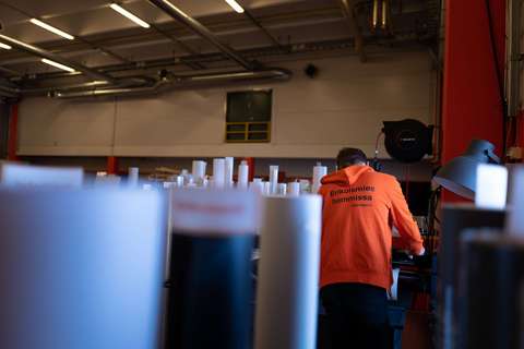 En man iklädd en orange huvtröja står och arbetar bland oskarpa, rörformade föremål. På huvtröjans baksida står det ”Erikoismies hommissa – Brändäämö”.