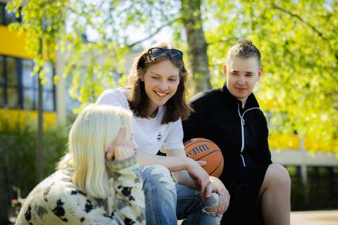 Three young people are sitting outside, two are looking at the third. In the background, there is a building and a birch tree.
