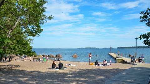 The picture shows Haukilahti beach. People are sitting on the sand and some people are standing on the pier. The sky is blue and the weather is sunny.
