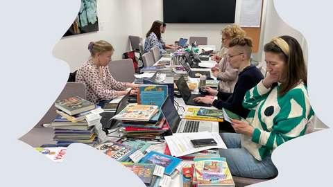 Opettajia ja kirjastolaisia työskentelemässä pitkän pöydän ääressä. Pöydällä on pinoissa paljon uusia lasten- ja nuortenkirjoja.