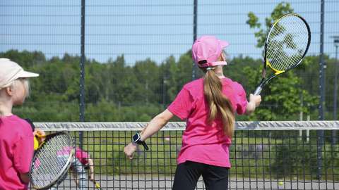 Kuvassa kaksi lasta pelaamassa tennistä ulkona. Taustalla näkyy vihreä luonto. Lapsilla on lippalakit päässä.