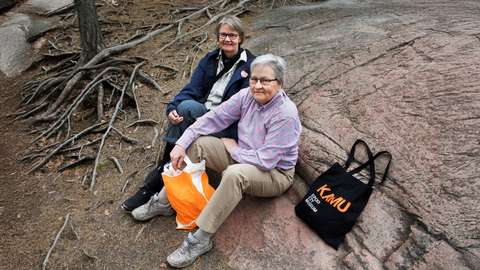 Sateenkaarisenioreiden Anne ja Päivi istuvat kalliolla ja katsovat ylös kameraan.